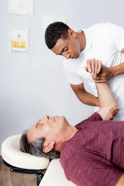 African american chiropractor correcting arm of middle aged man in clinic — Stock Photo