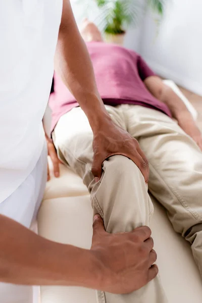 Vista ritagliata del chiropratico afroamericano che lavora con il ginocchio dell'uomo di mezza età in clinica — Foto stock