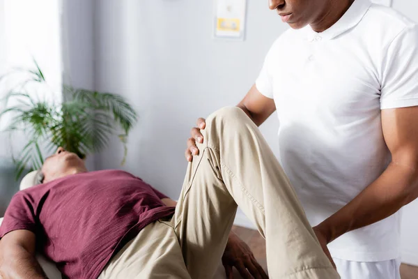 Chiropraticien afro-américain corrigeant le genou d'un homme d'âge moyen en clinique — Photo de stock