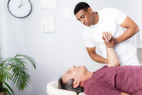 African american chiropractor correcting arm of middle aged man on massage table — Stock Photo