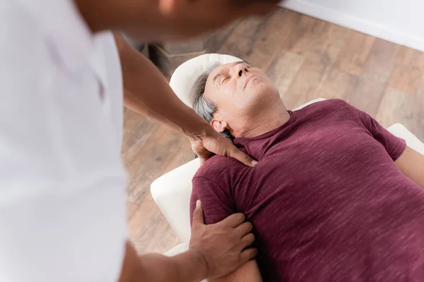 Desfocado afro-americano quiroprático massagem braço de homem maduro na mesa de massagem — Fotografia de Stock