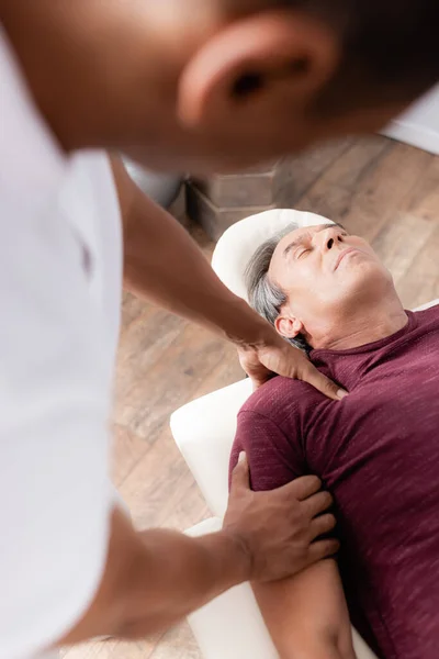 Borrosa afroamericana quiropráctico masaje brazo de hombre de mediana edad en mesa de masaje - foto de stock