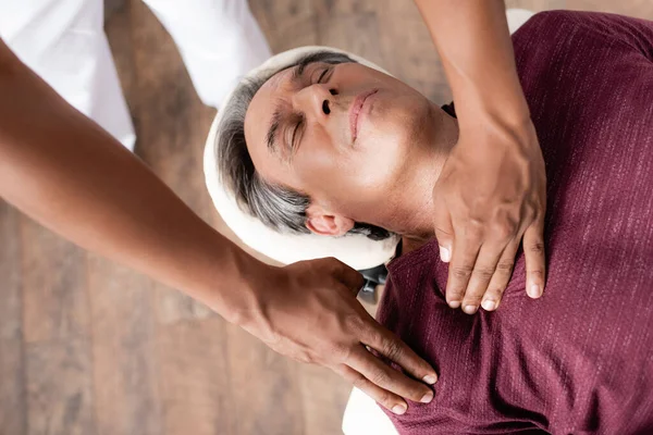 Vista de ángulo alto de masajista afroamericano masajeando hombre de mediana edad con los ojos cerrados en la mesa de masaje - foto de stock