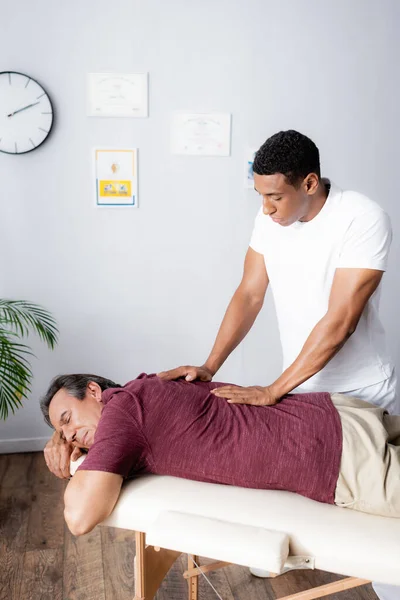 Massagista afro-americano massageando costas de homem de meia-idade na mesa de massagem — Fotografia de Stock