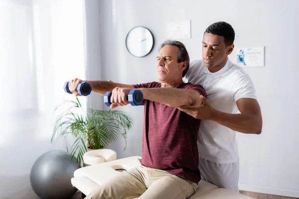 Middle aged man exercising with dumbbells on massage table near african american physiotherapist — Stock Photo