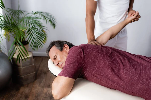 African american physiotherapist correcting arm of middle aged man on massage table — Stock Photo