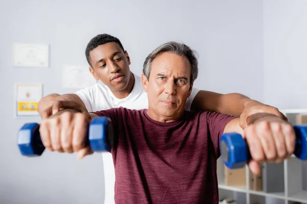 Fisioterapeuta afroamericano ayudando a un hombre de mediana edad a hacer ejercicio con pesas en primer plano borroso - foto de stock