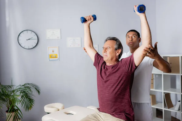 Hombre de mediana edad haciendo ejercicio con pesas cerca de fisioterapeuta afroamericano - foto de stock