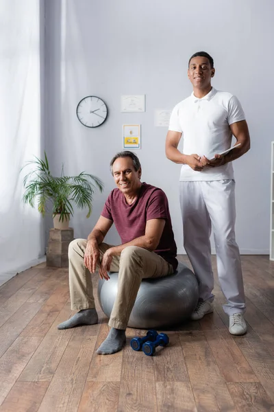 Feliz hombre de mediana edad sentado en la pelota de fitness cerca de afroamericano fisioterapeuta con tableta digital - foto de stock