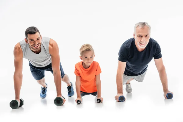 Alegre chico con abuelo y papá haciendo flexiones ejercicio con mancuernas en blanco - foto de stock