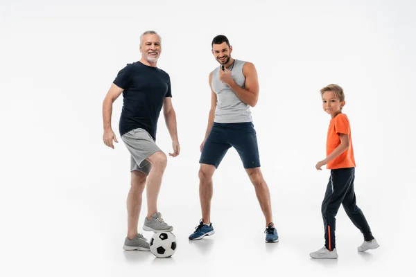 Cheerful man whistling near boy and grandfather stepping on soccer ball on white — Stock Photo