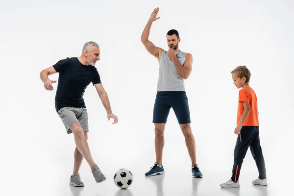 Homme en vêtements de sport sifflant près de grand-père jouer au football avec petit-fils sur blanc — Photo de stock