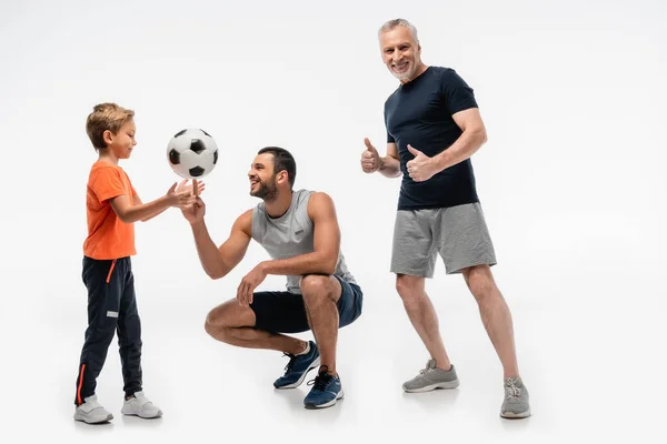 Fröhlicher Opa zeigt Daumen hoch in der Nähe von Vater und Sohn beim Spielen mit Fußball auf Weiß — Stockfoto