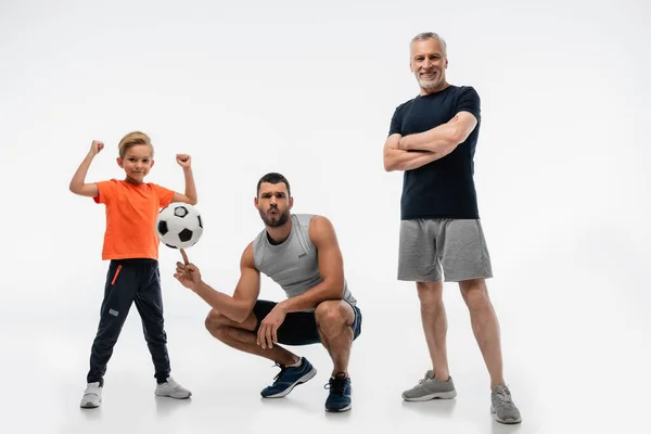 Man in sportswear whistling while playing with soccer ball near boy and grandfather on white — Stock Photo