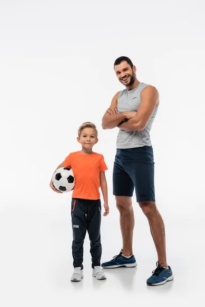 Hombre con los brazos cruzados sonriendo a la cámara cerca de hijo con pelota de fútbol en blanco - foto de stock