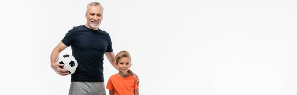 Cheerful man holding hand on shoulder of grandson while holding soccer ball isolated on white, banner — Stock Photo