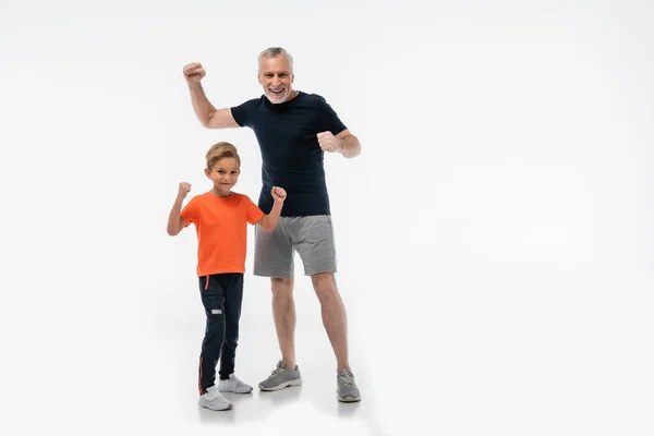 Excité grand-père avec petit-fils en vêtements de sport montrant geste de victoire sur blanc — Photo de stock