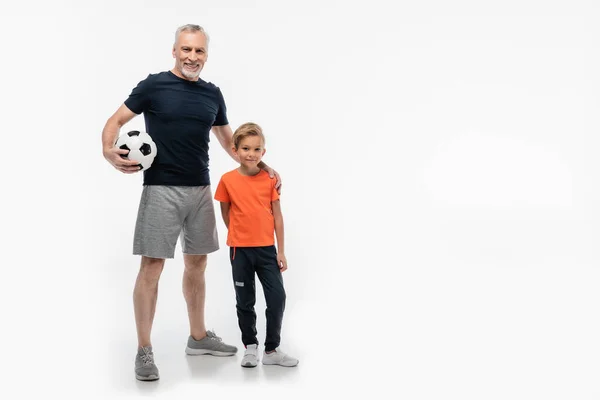 Homme heureux en vêtements de sport tenant la main sur l'épaule du petit-fils tout en tenant ballon de football sur blanc — Photo de stock