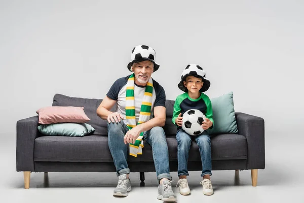 Smiling grandfather and grandson watching football match while sitting on sofa in fan hats on grey — Stock Photo