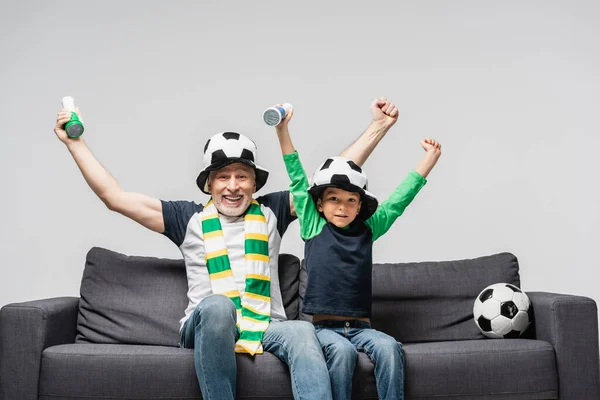 Uomo eccitato con nipote che mostra il gesto di vittoria mentre guarda il campionato di calcio in cappelli dei tifosi isolati sul grigio — Foto stock