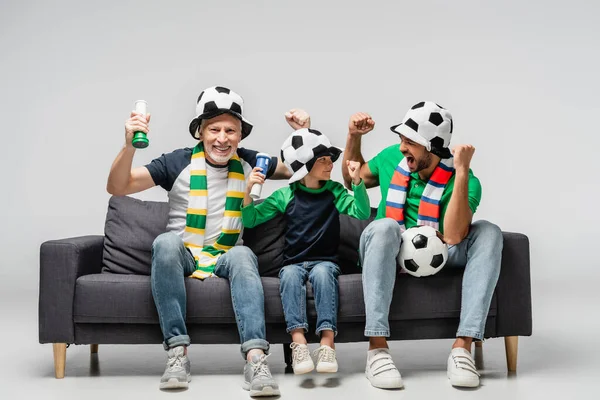 Ragazzo eccitato con nonno e papà mostrando vincere gesto mentre si guarda la partita di calcio in cappelli da tifoso su grigio — Foto stock