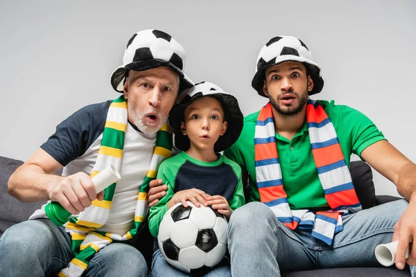 Chico sorprendido con pelota de fútbol viendo campeonato de fútbol junto con el padre y el abuelo aislado en gris - foto de stock