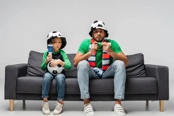 Tendu papa et fils dans les chapeaux de ventilateur regarder match de football sur gris — Photo de stock