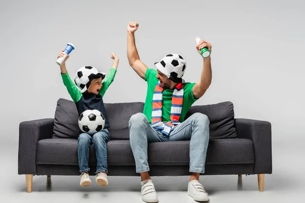 Excité père et fils en chapeaux de ventilateur se regardant tout en montrant geste de victoire sur gris — Photo de stock