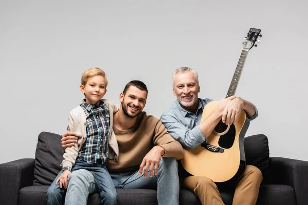 Glücklicher reifer Mann mit Akustikgitarre sitzt auf Sofa neben Sohn und Enkel isoliert auf grau — Stockfoto