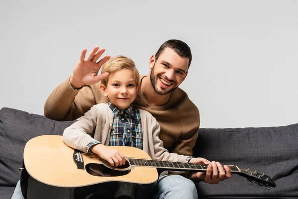 Веселый мальчик, играющий на акустической гитаре рядом с счастливым отцом, машущим рукой в камеру, изолированную на сером — стоковое фото
