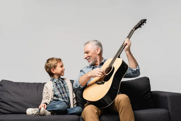 Abuelo feliz tocando la guitarra acústica cerca de nieto sonriente aislado en gris - foto de stock