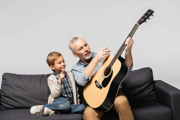 Mature man pointing with finger at acoustic guitar near grandson isolated on grey — Stock Photo