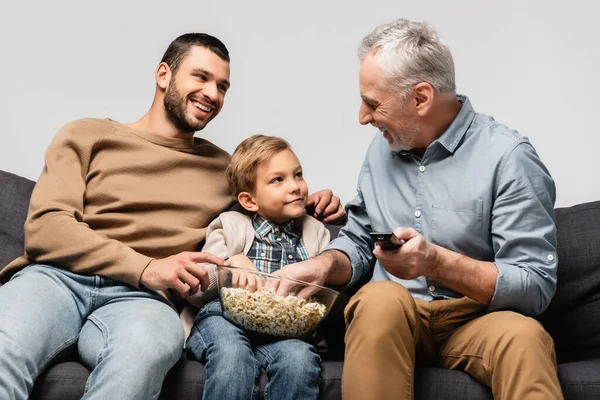 Happy mature man sitting on sofa with remote controller near son and grandson with bowl of popcorn isolated on grey — Stock Photo