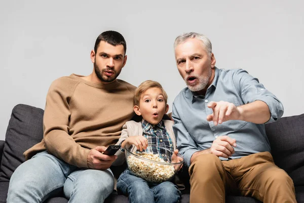 Homem surpreso apontando com o dedo enquanto assiste tv perto de filho e neto com tigela de pipoca isolada em cinza — Fotografia de Stock
