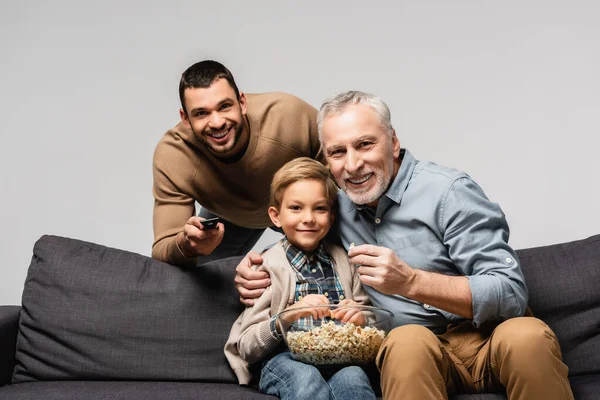 Happy man holding remote controller near father and son watching tv and eating popcorn isolated on grey — Stock Photo