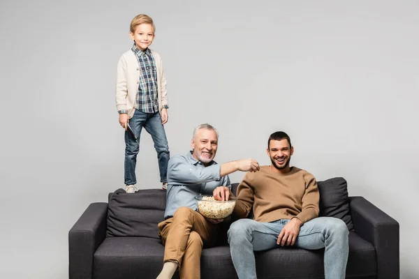 Smiling boy standing on sofa near dad and grandfather watching tv and eating popcorn isolated on grey — Stock Photo