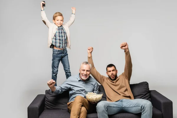 Homens animados com menino mostrando gesto de vitória enquanto assiste tv no sofá isolado no cinza — Fotografia de Stock