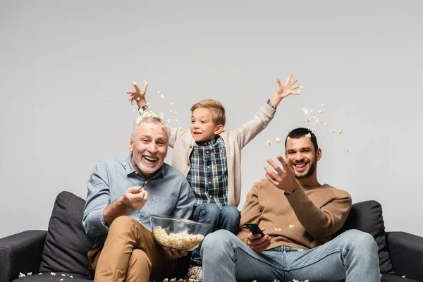 Excited boy with dad and grandfather throwing popcorn while watching tv isolated on grey — Stock Photo