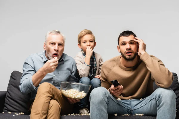 Astonished man holding bowl with popcorn while watching tv together with son and grandson isolated on grey — Stock Photo