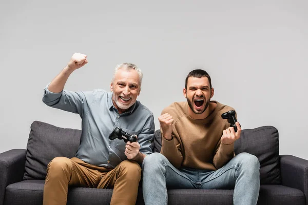 QUIIV, UCRÂNIA - NOVEMBRO 17, 2020: pai animado com o filho adulto segurando joysticks e mostrando gesto de vitória isolado em cinza — Fotografia de Stock