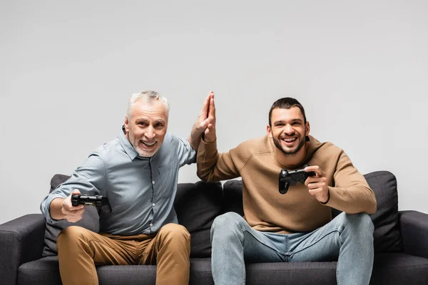 KYIV, UCRANIA - 17 DE NOVIEMBRE DE 2020: padre e hijo emocionados dando cinco altos mientras están sentados en el sofá con joysticks aislados en gris - foto de stock