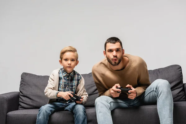 KYIV, UCRANIA - 17 de noviembre de 2020: padre e hijo concentrados jugando videojuegos en un sofá aislado en gris - foto de stock