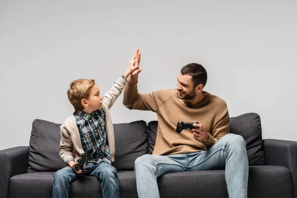 QUIIV, UCRÂNIA - NOVEMBRO 17, 2020: pai e filho alegres segurando joysticks e dando alta cinco isolados em cinza — Fotografia de Stock