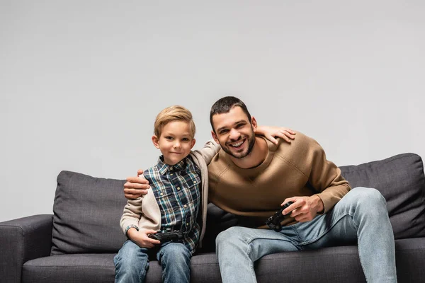 KYIV, UCRANIA - 17 DE NOVIEMBRE DE 2020: feliz padre e hijo abrazando mientras sostiene joysticks aislados en gris - foto de stock