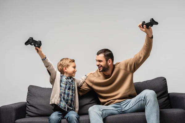 KYIV, UKRAINE - 17 NOVEMBRE 2020 : le père et le fils joyeux montrant geste de victoire avec des joysticks isolés sur gris — Photo de stock