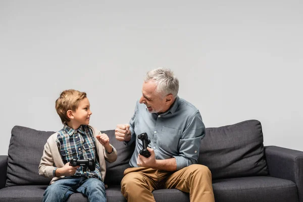 KYIV, UCRANIA - 17 DE NOVIEMBRE DE 2020: abuelo feliz y nieto haciendo golpe de puño mientras sostiene joysticks aislados en gris - foto de stock