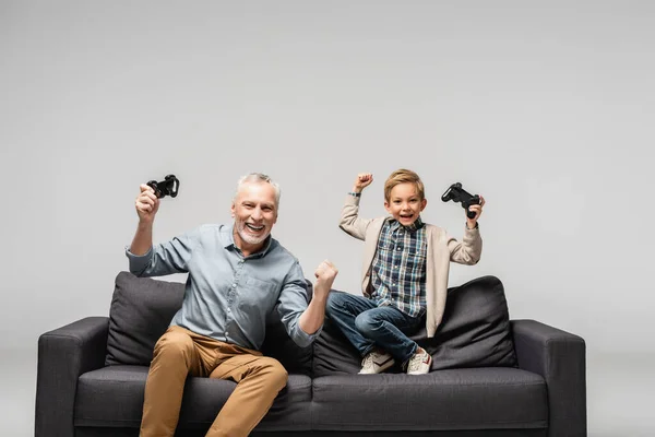 KYIV, UCRANIA - 17 de noviembre de 2020: niño emocionado con el abuelo mostrando gesto de victoria con joysticks aislados en gris - foto de stock