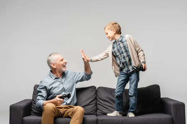 KYIV, UKRAINE - NOVEMBER 17, 2020: happy mature man giving high five to grandson while holding joystick isolated on grey — Stock Photo