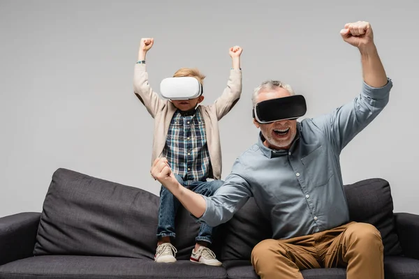 Hombre excitado con nieto en auriculares vr mostrando gesto de victoria aislado en gris - foto de stock