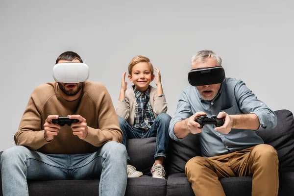 KYIV, UKRAINE - NOVEMBER 17, 2020: cheerful boy near concentrated grandfather and dad playing video game with joysticks isolated on grey — Stock Photo